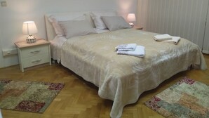 A neatly made bed with a beige bedspread, two bedside lamps, and a small bedside table with a lamp. There are two decorative rugs on the wooden floor.