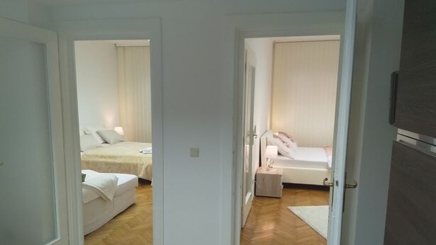A hotel room with a wooden floor, a bed with white bedding, a nightstand with a lamp, and a door leading to another room.