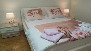 A neatly made bed with floral pillows and a pink blanket, two bedside lamps, and a wooden floor.