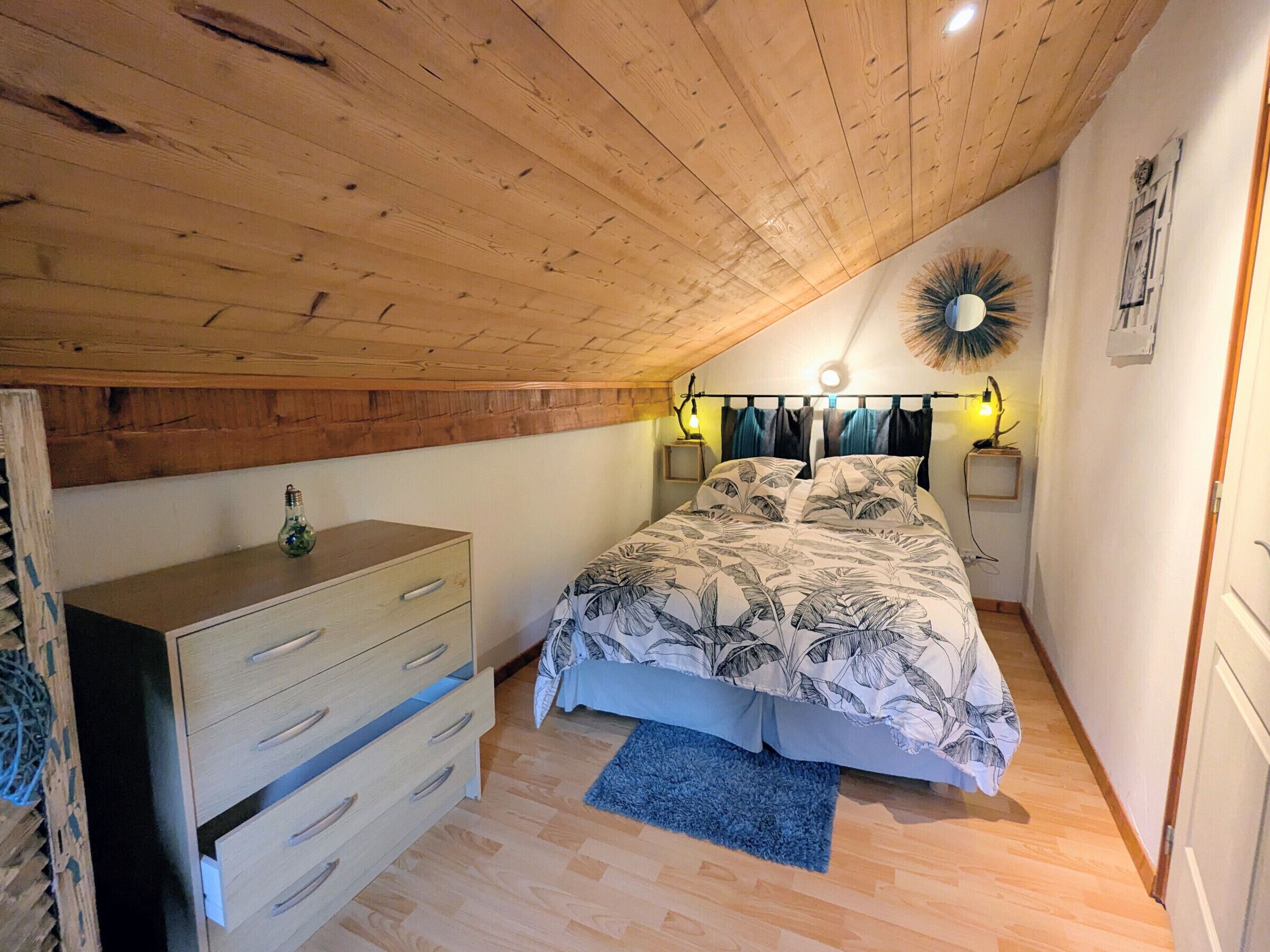 A bedroom with a sloped ceiling, a bed with a leaf-patterned bedspread, a wooden dresser, and two bedside lamps.