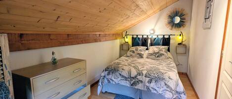 A bedroom with a sloped ceiling, a bed with a leaf-patterned bedspread, a wooden dresser, and two bedside lamps.