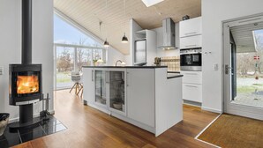 A modern kitchen with a central island, a wood-burning stove, and a large window overlooking a garden.