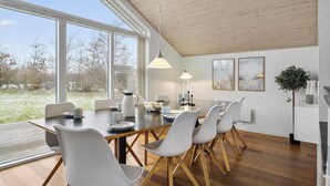 A dining area with a large table, white chairs, and a view of a natural landscape through large windows.