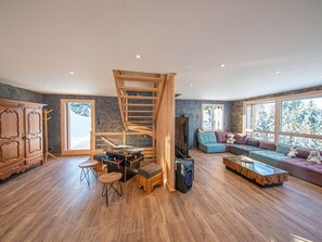 A spacious living room with a wooden staircase, a large window with a view of trees, a sofa set, and a wooden cabinet.