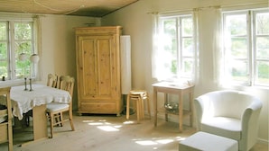 A room with a wooden dining table, chairs, a white armchair, a small table with a lamp, and a large wooden cabinet.