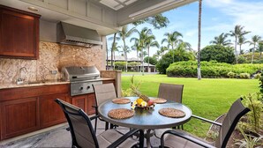 An outdoor kitchen with a grill, a round table with chairs, and a view of a well-maintained lawn and palm trees.