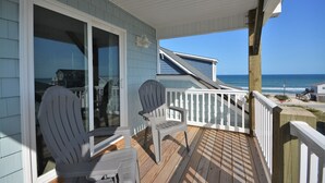 A wooden deck with two Adirondack chairs, a glass door leading to a room, and a view of the ocean. - Experience coastal views at Topsail Turvy, a spacious 4-bedroom home in North Topsail Beach. (North Topsail Beach)
