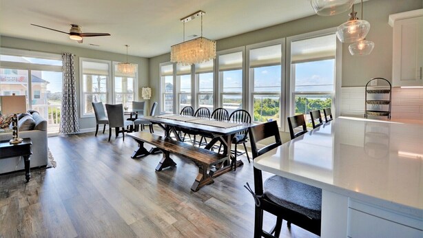 A spacious dining area with a long table and chairs, large windows, and a ceiling fan. - Experience coastal views at Topsail Turvy, a spacious 4-bedroom home in North Topsail Beach. (North Topsail Beach)