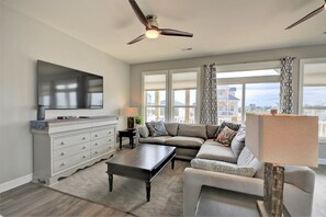 A modern living room with a sectional sofa, a flat-screen TV, a ceiling fan, and large windows offering a city view.
