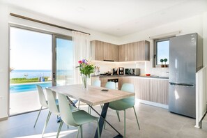 A modern kitchen with a stainless steel refrigerator, a dining area with a table and chairs, and a view of the ocean through large sliding glass doors.