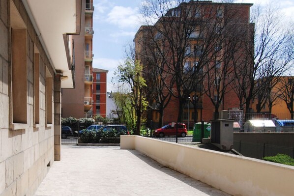 A narrow urban street with a row of multi-story buildings, a paved walkway, and parked cars.
