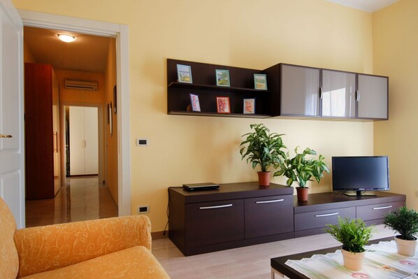 A living room with a sofa, a TV, a cabinet with shelves, and potted plants.