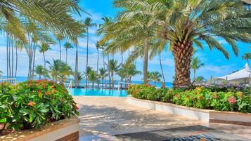 Zona de piscina en un resort con palmeras, cielo azul despejado y vista al océano.