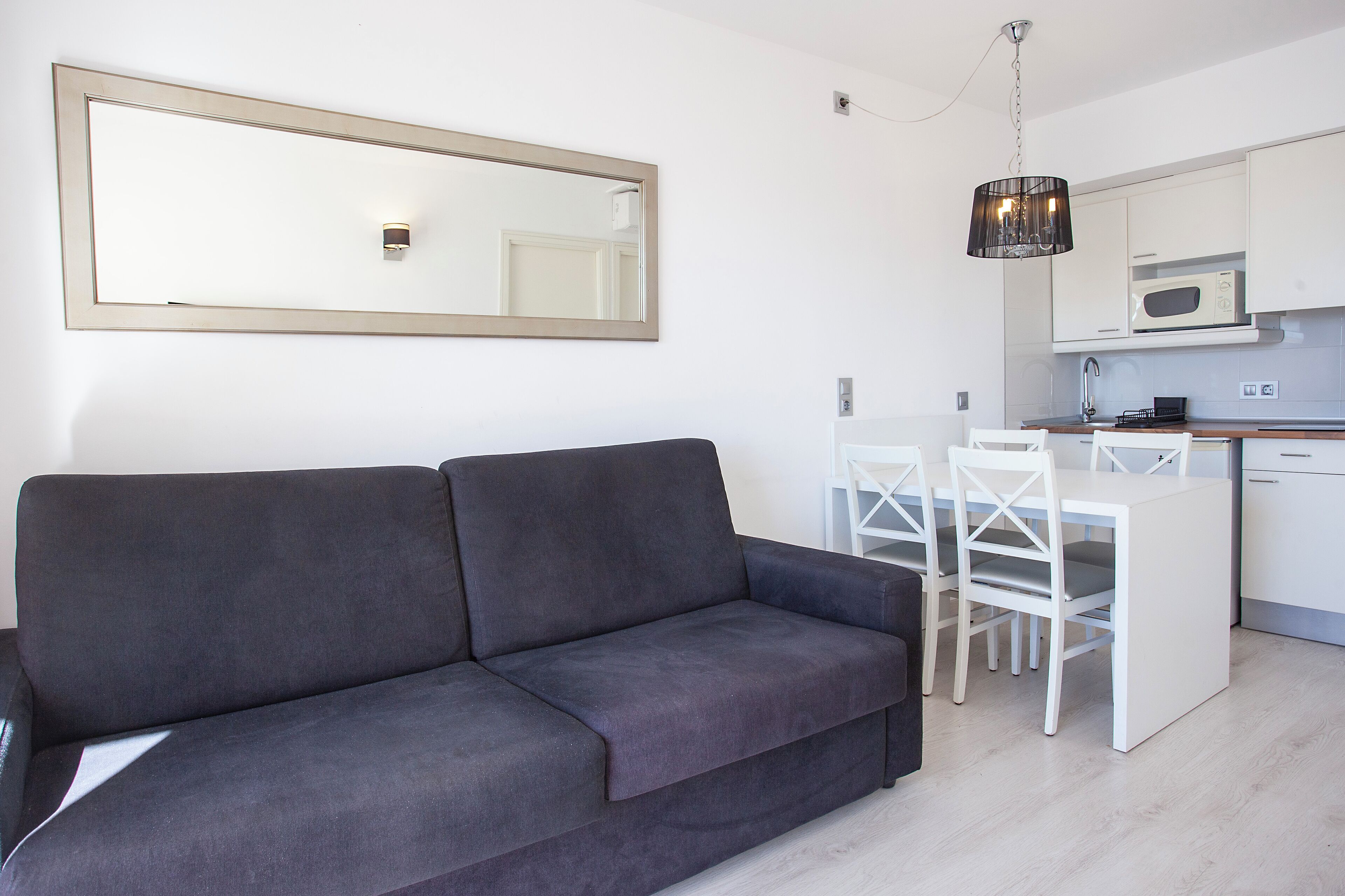 A modern living room with a grey sofa, a dining table with white chairs, a kitchen area with white cabinets, and a large mirror on the wall.