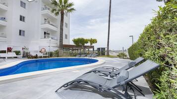 Una piscina con sillas de descanso, un edificio blanco con balcones y una palmera.