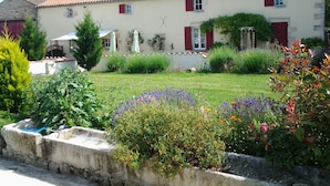 A two-story building with a tiled roof, red shutters, and a garden with various plants and flowers. - 6 bedroom Traditional Farmhouse, large pool, indoor & outdoor games areas. (Rives-d'Autise)