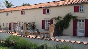 A building with a tiled roof, red shutters, and a garden area with a wooden fence and plants. - 6 bedroom Traditional Farmhouse, large pool, indoor & outdoor games areas. (Rives-d'Autise)