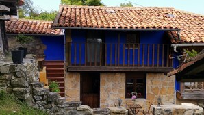 A two-story stone house with a tiled roof, a balcony with wooden railings, and a stone wall surrounding the property.