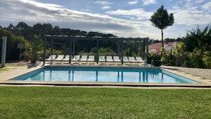 Une piscine entourée de chaises longues sous une pergola, le tout baigné de verdure et surplombé par un ciel dégagé.