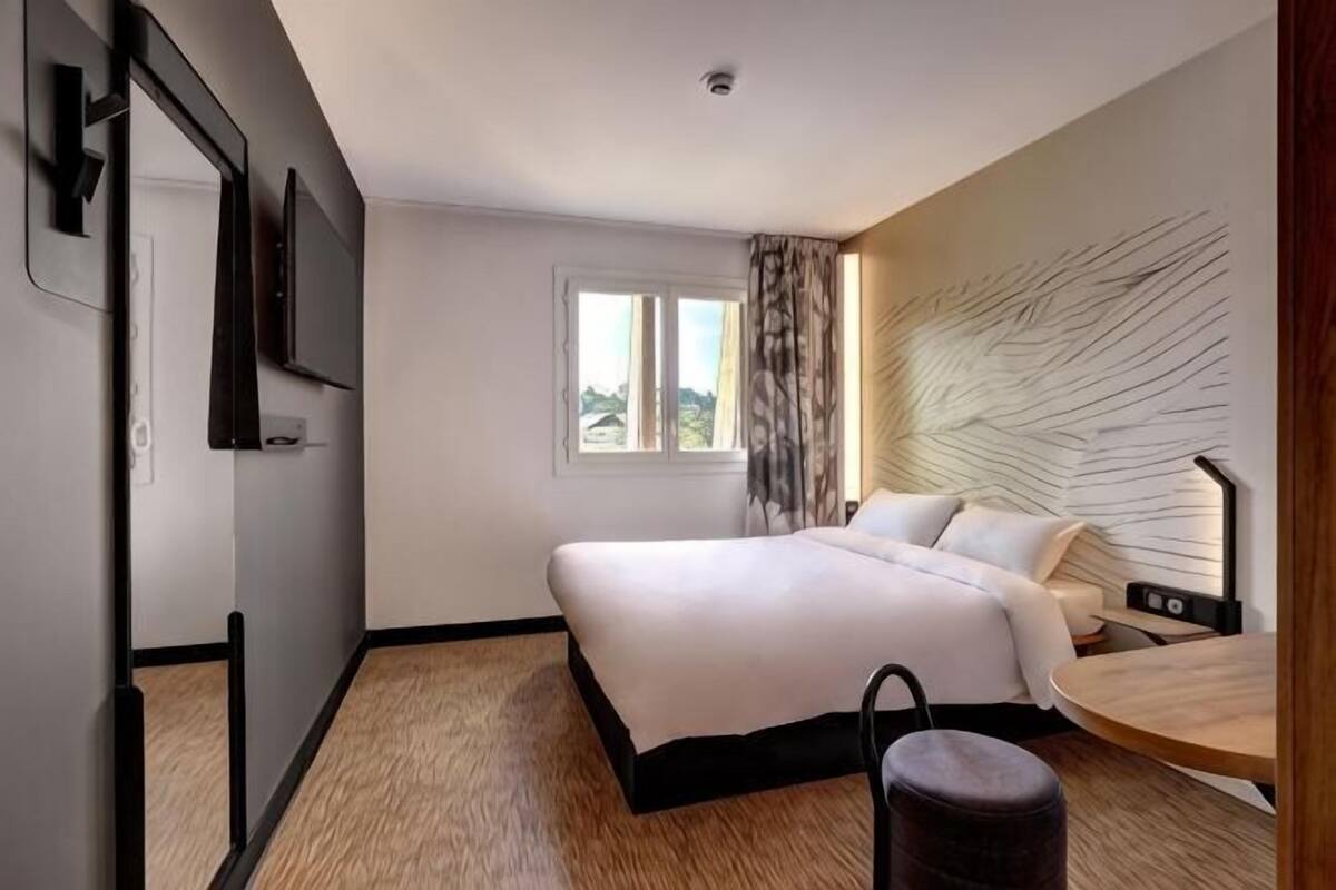 Ein modernes Hotelzimmer mit Bett, Schreibtisch, Stuhl, Fenster und einem am Wand befestigten Fernseher.
