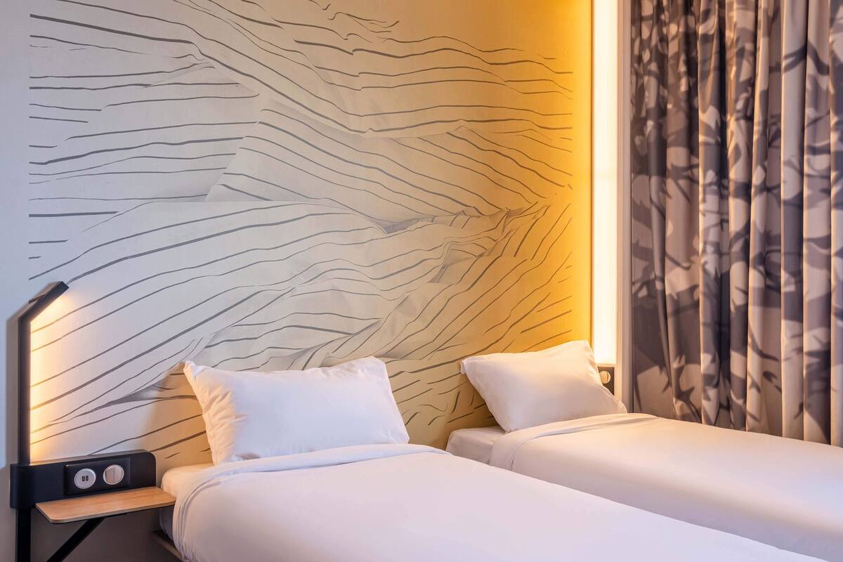 Ein Hotelzimmer mit zwei Betten, einem Kopfteil mit wellenförmigem Muster, einem Nachttisch mit Radio und einem Fenster mit gemusterten Vorhängen.