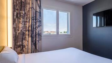 Ein Hotelzimmer mit Bett, Fenster mit Blick auf Gebäude und einem an der Wand befestigten Flachbildfernseher.