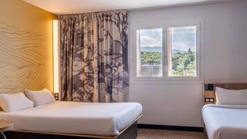 Quarto de hotel com cama, uma cadeira, janela com vista para áreas verdes e luminária na parede.