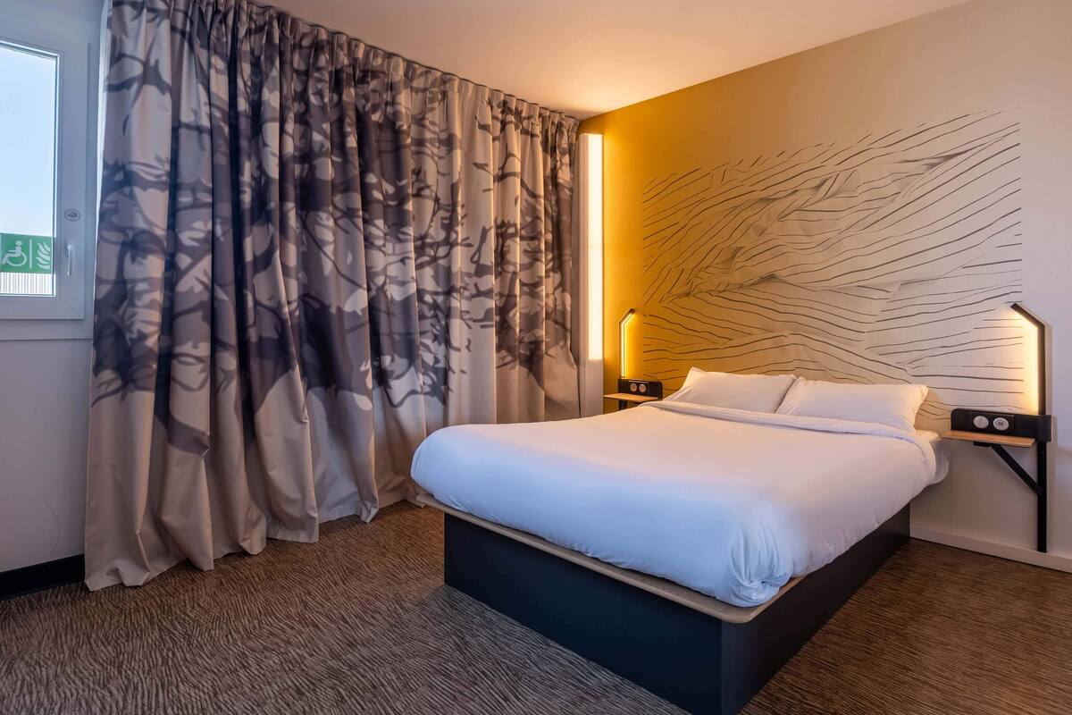 Ein Hotelzimmer mit Bett, Vorhängen, einem Wandbild, einem Nachttisch und einer Lampe.