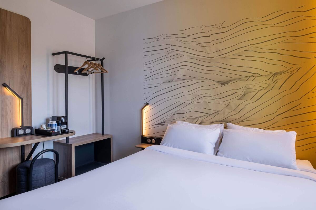 Ein Hotelzimmer mit einem Bett, einem Schreibtisch mit Stuhl, einem Kleiderständer und einer Wand mit strukturierter Gestaltung.