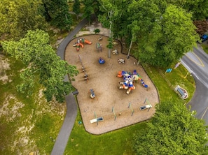 Explore a playground surrounded by lush greenery, featuring slides, swings, and climbing frames on a sandy surface.