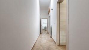 Un corridoio stretto con pavimento piastrellato, pareti bianche e una porta in legno sulla destra.