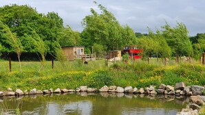 Un estanque rodeado de rocas y vegetación, una construcción de madera y un autobús rojo.