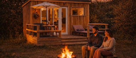 A wooden cabin with a deck, a fire pit, and a couple sitting outside.