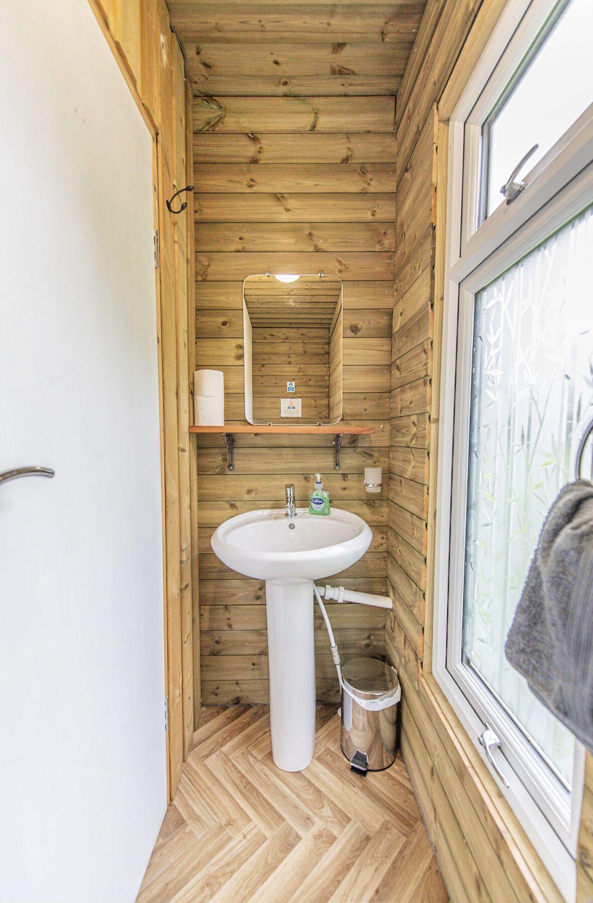 Ein kleines Badezimmer aus Holz mit Waschbecken in Weiß, einem Spiegel und einem Fenster.
