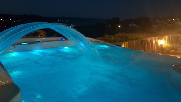 Ein Whirlpool mit Wasserfall, umgeben von einem Holzzaun und beleuchtet mit stimmungsvoller Beleuchtung.