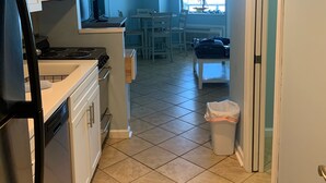 A compact kitchen with white cabinets, a black refrigerator, and a stove. There is a dining area with a table and chairs, and a trash can in the foreground. - Beautiful Ocean view corner unit (Myrtle Beach)