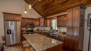 A kitchen with wooden cabinets, a stainless steel refrigerator, a granite countertop, and bar stools. - Pristine Cabin in Heart of Boone (Vilas)
