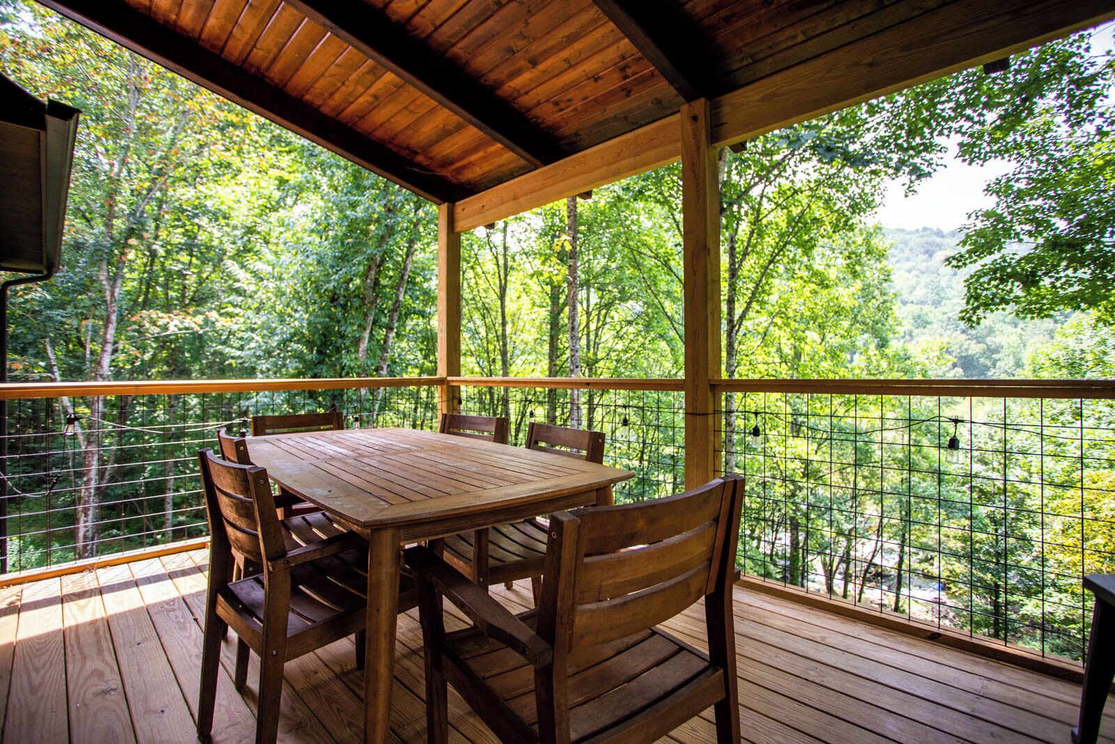 Eine Holzdielen-Terrasse mit Tisch und Stühlen, umgeben von einem Metallzaun, mit Blick auf ein bewaldetes Gebiet.