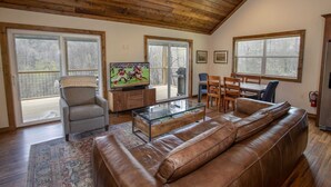 A spacious living room with a wooden ceiling, large windows, a leather sofa, a glass coffee table, and a television. - Pristine Cabin in Heart of Boone (Vilas)