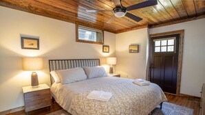 A bedroom with a bed, two bedside tables with lamps, a ceiling fan, wooden flooring, a door, and framed pictures on the walls. - Pristine Cabin in Heart of Boone (Vilas)