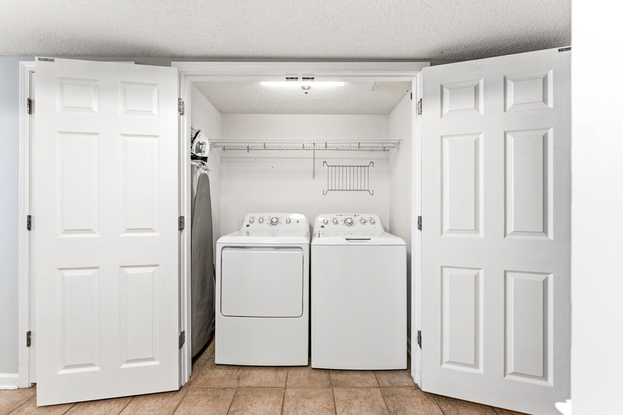 A laundry room with two white front-loading washers and dryers, a metal drying rack, and a ceiling light.