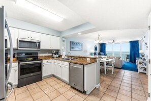A modern kitchen with white cabinets, a microwave, stove, and dishwasher. There's a dining area with a table and chairs, and a view of the ocean through large windows. - Luxury oceanfront condo - Indoor/Outdoor Pools + Free Attraction Tickets! (North Myrtle Beach)