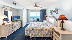 A hotel room with a bed, a TV, a ceiling fan, a balcony with a view of the ocean, and a desk with a lamp.