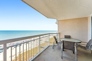A balcony with a table and chairs, overlooking a beach with people and umbrellas. - Luxury oceanfront condo - Indoor/Outdoor Pools + Free Attraction Tickets! (North Myrtle Beach)
