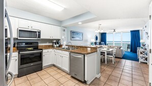 A modern kitchen with white cabinets, a microwave, stove, and dishwasher. There's a dining area with a table and chairs, and a view of the ocean through large windows.