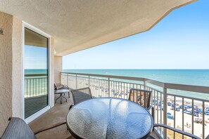 A balcony with a glass table and chairs, overlooking a beach with umbrellas and people. - Luxury oceanfront condo - Indoor/Outdoor Pools + Free Attraction Tickets! (North Myrtle Beach)