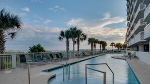 A swimming pool with lounge chairs and palm trees, surrounded by a fence and a building with balconies.