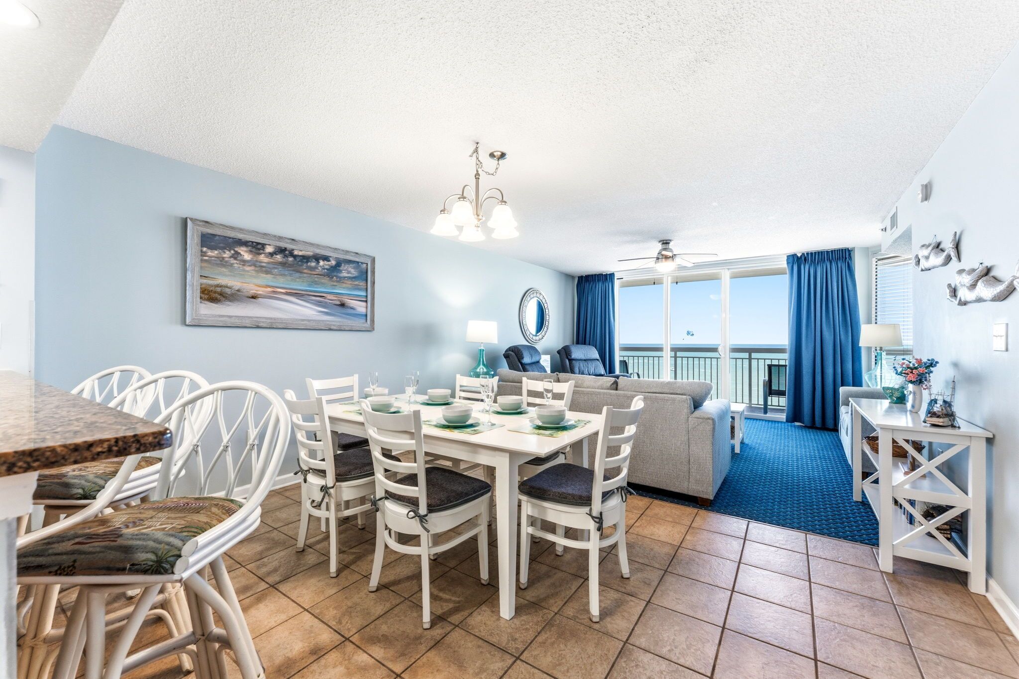 Un coin repas avec une table dressée pour quatre personnes, des chaises blanches, un ventilateur de plafond et une vue sur l’extérieur.