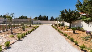 A gravel pathway leading to a building with a white wall and a garden with small plants and trees. - 1890 Bilo Conchiglia Blu (Porto Cesareo)