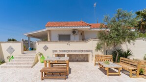 A house with a tiled roof, a covered patio with a table and benches, and a decorative wall. - 1890 Bilo Conchiglia Blu (Porto Cesareo)